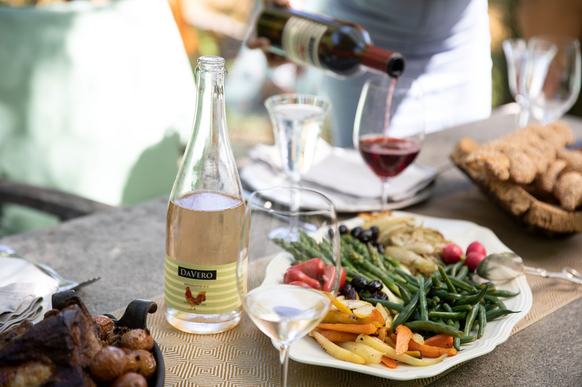 This image shows an outdoor table setting featuring a variety of prepared dishes, including roasted vegetables, asparagus, green beans, and a platter of crusty bread. A bottle of DaVero wine stands prominently alongside wine glasses, capturing a warm and inviting al fresco dining atmosphere. The scene suggests a relaxed, high-quality outdoor entertaining space suitable for social gatherings.