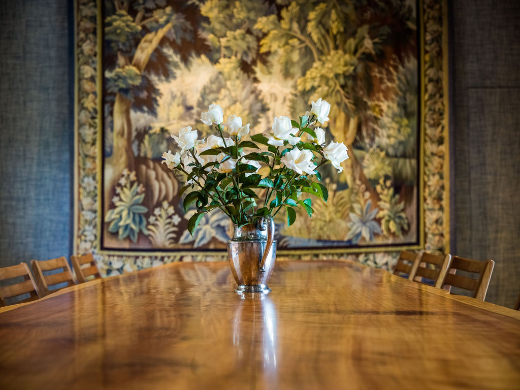 This interior shot highlights a refined dining room centered around a long, polished wooden table. A metallic pitcher holding white roses serves as a sophisticated focal point, positioned in front of a decorative, nature-themed wall tapestry. The space is framed by wooden dining chairs, suggesting an elegant and inviting atmosphere for formal dining.