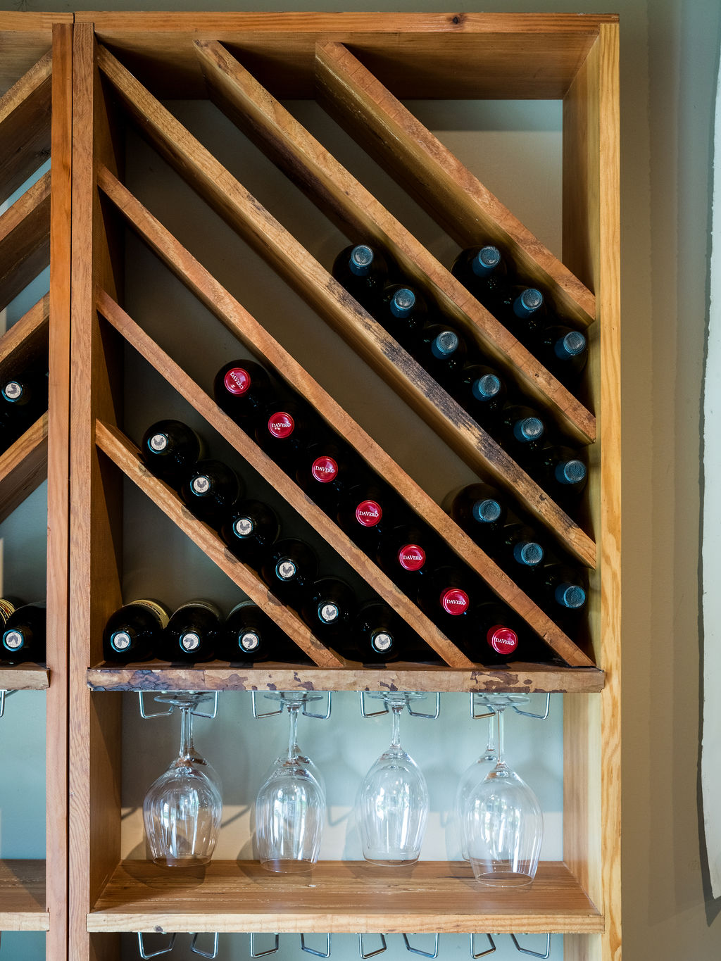 A custom-built wooden wine rack provides elegant storage, featuring slanted shelving that displays multiple DaVero wine bottles. Below the rack, a row of stemware hangs neatly from integrated racks, creating a sophisticated and functional display area. This organized storage solution adds a touch of luxury and charm suitable for a home wine cellar or tasting area.