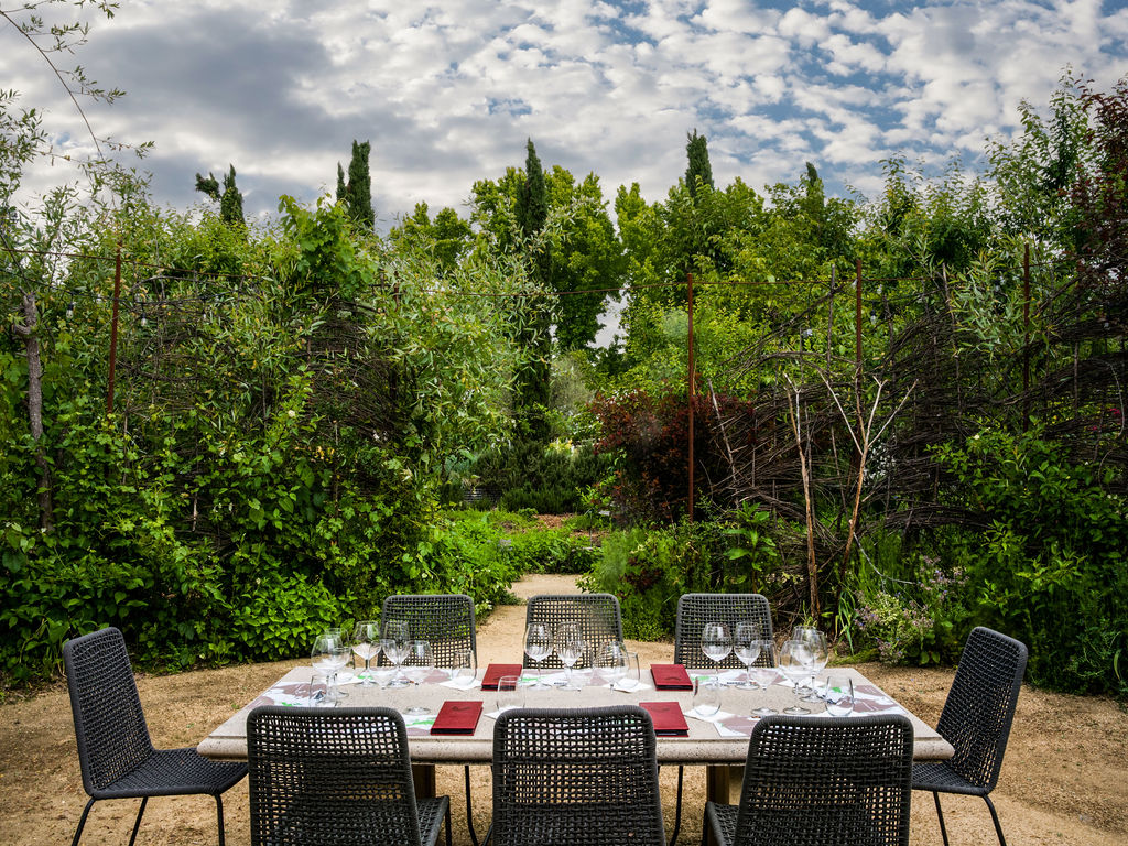 This outdoor patio space features a modern dining set with dark woven chairs and a long stone table, perfectly situated for alfresco dining. Surrounded by lush greenery and dense landscaping, the area offers a serene and private atmosphere. The setup includes multiple wine glasses and place settings, suggesting a high-end, sophisticated garden dining environment ideal for entertaining.