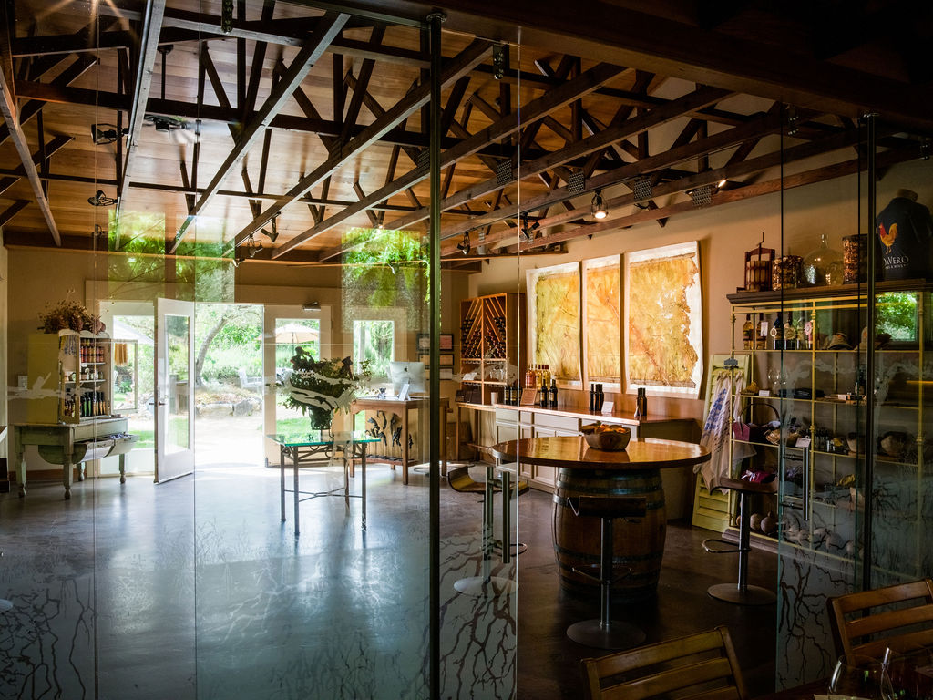 This image showcases a charming, rustic-style winery tasting room or boutique retail space. The interior features exposed wooden trusses on the ceiling, a central wine barrel table with bar stool seating, and open shelving displays for wine-related merchandise, all creating a welcoming, artisanal atmosphere that opens up to an outdoor garden area.