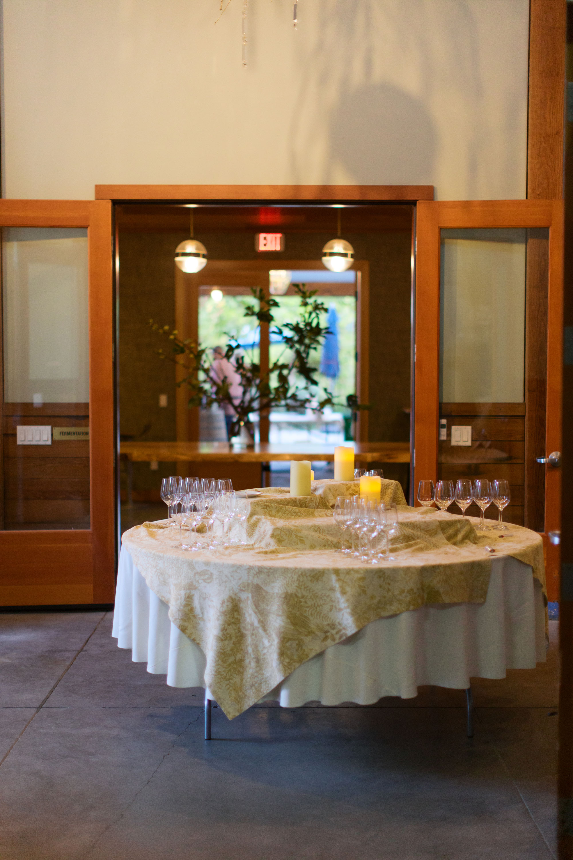 This image features a welcoming entryway into a communal or event space, highlighted by a set of double wooden doors leading into a room arranged for a gathering. A circular table with a textured runner, multiple wine glasses, and illuminated pillar candles creates an elegant and inviting atmosphere. The view through the open doors into the adjacent room, which has large windows and garden scenery, suggests a versatile, well-maintained event venue or clubhouse.