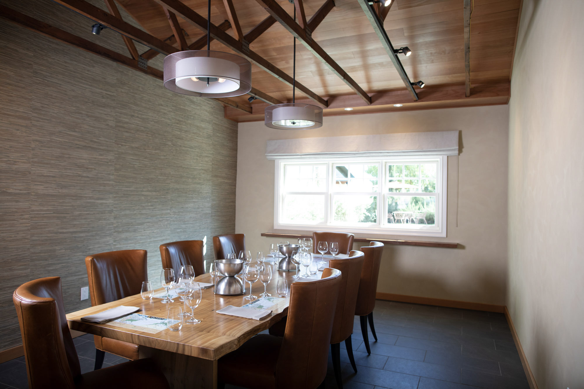 This elegant dining room features a large, rustic live-edge wood table surrounded by high-backed tan leather chairs, perfect for formal entertaining. The space is anchored by two contemporary drum pendant lights and highlighted by a sophisticated textured accent wall and an exposed wood-beam ceiling. A large window brings in natural light, overlooking the serene exterior landscape.