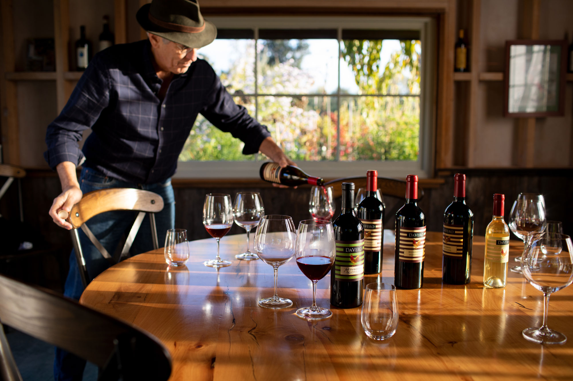 This warm and inviting dining space features a large, rustic circular wood table set for wine tasting. An individual is shown pouring red wine into a glass, highlighting the room's function as an elegant venue for entertaining. Large, multi-pane windows bathe the interior in soft, natural light, creating an atmosphere of sophistication and comfort.
