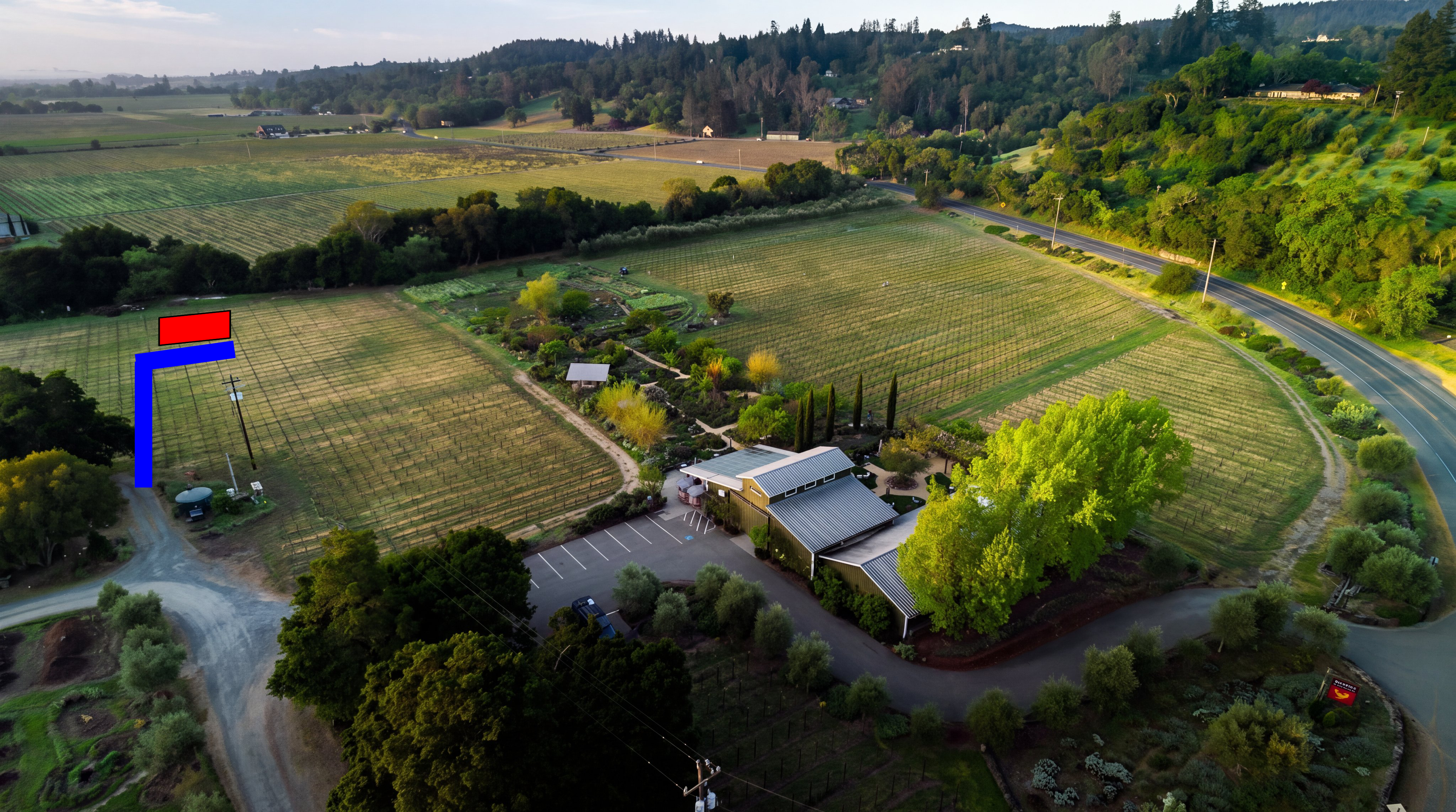 This stunning aerial view captures a picturesque vineyard estate, showcasing rows of well-maintained vines, lush landscaped gardens, and a distinctive barn-style building with a metal roof. The property is nestled within a serene, rolling landscape, offering a perfect blend of rural charm and agricultural appeal. With its paved driveway, ample parking, and peaceful surroundings, this estate presents a highly desirable and scenic setting.