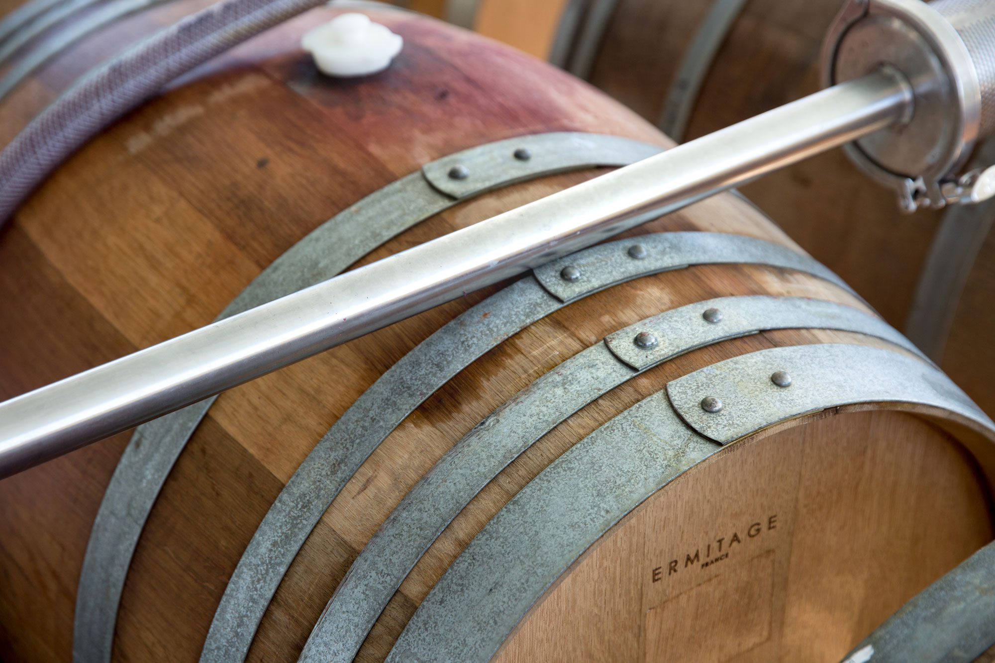 This close-up image features a wooden wine barrel with metal hoops, branded with the name 'ERMITAGE FRANCE'. A metal piping system or racking fixture is positioned across the top, indicating a functional winemaking or storage environment. The shot captures the authentic, industrial, and artisanal quality of professional wine aging facilities.