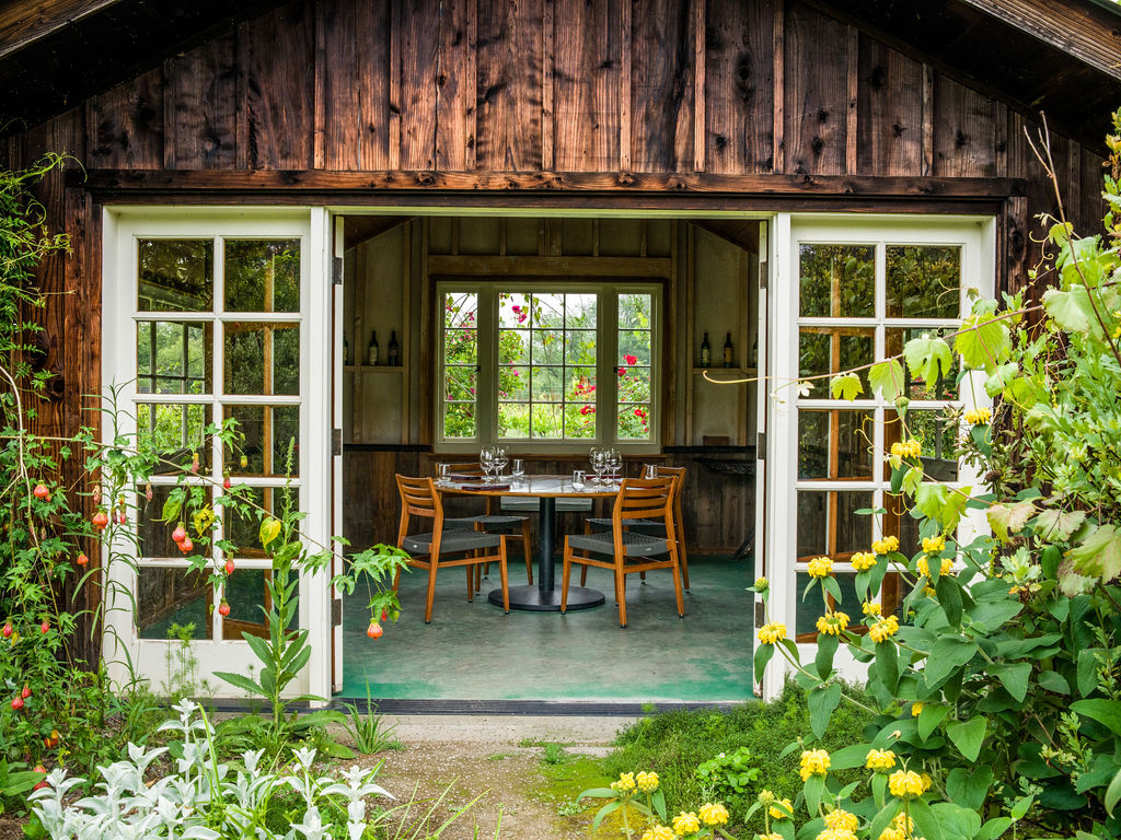 This charming, rustic dining space is set within a garden structure featuring weathered wood siding and French-style doors that open directly to the outdoors. The interior is minimalist with a circular dining table, wooden chairs, and large windows that frame the lush surrounding garden, creating an intimate, serene atmosphere. It serves as a unique, nature-integrated dining retreat.