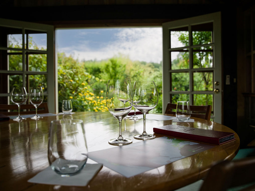 This dining room space features a polished wooden table set with assorted wine glasses and menus, creating an inviting atmosphere for wine tastings or formal dinners. The room is framed by large French doors that provide a seamless view of a lush, vibrant green garden. The natural light flooding in from the outdoor setting enhances the warm, elegant aesthetic of the interior.