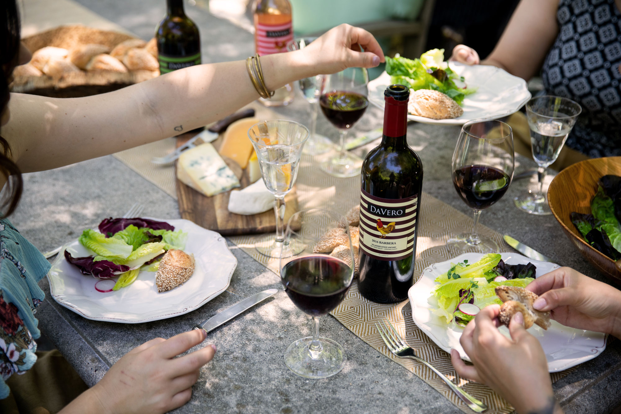 This image captures a close-up, elevated view of an outdoor dining setting featuring people enjoying a meal with wine and artisanal cheeses. The rustic table is arranged with wine glasses, plates of salad, and a bottle of DaVero wine, suggesting an inviting and social lifestyle atmosphere. The scene emphasizes an elegant outdoor entertaining space suitable for high-end residential marketing.