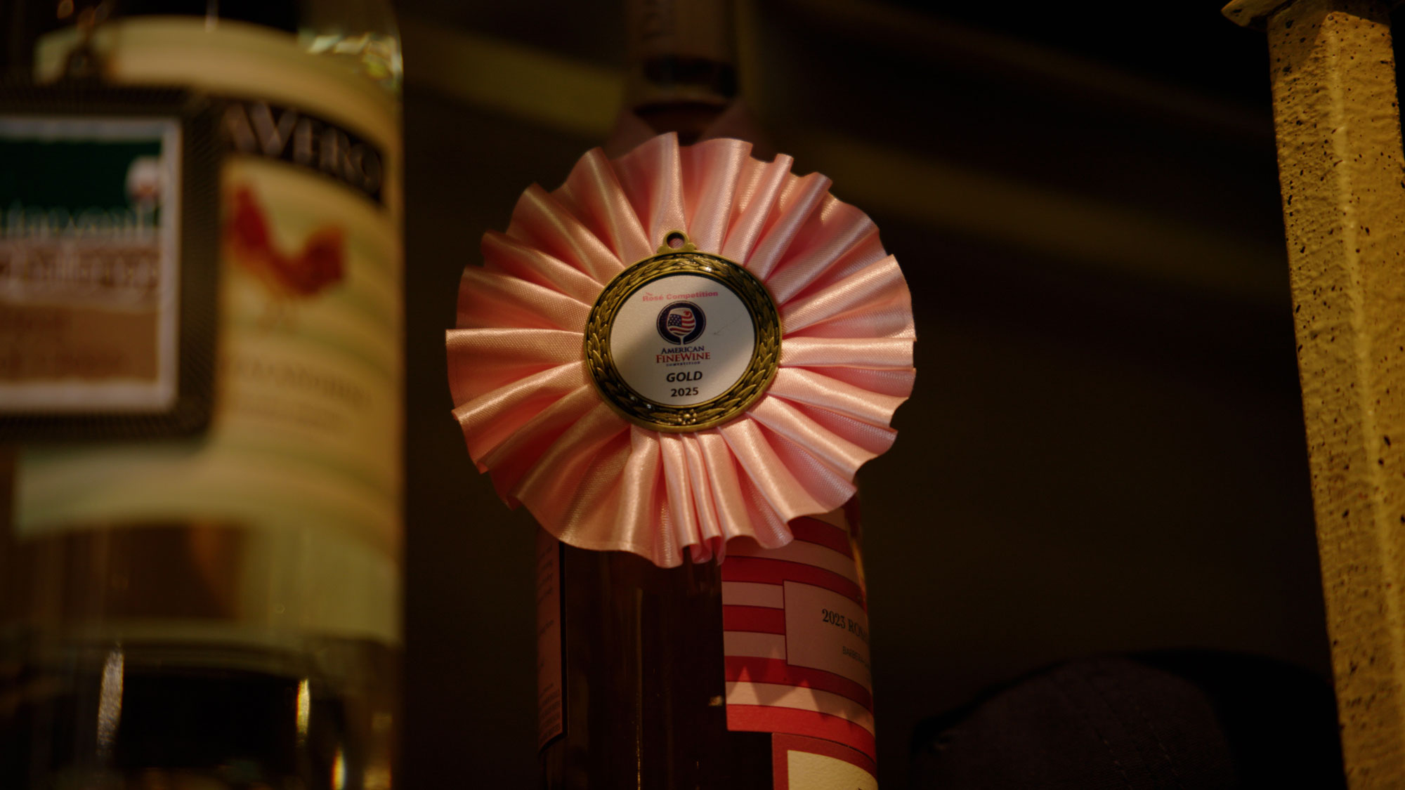 This image focuses on a wine bottle adorned with a 'Gold' award ribbon from the 2025 American Fine Wine Rosé Competition. The scene suggests a curated wine collection, highlighting high-quality selections that would appeal to wine enthusiasts. The lighting creates an intimate and sophisticated atmosphere suitable for a home wine cellar display.