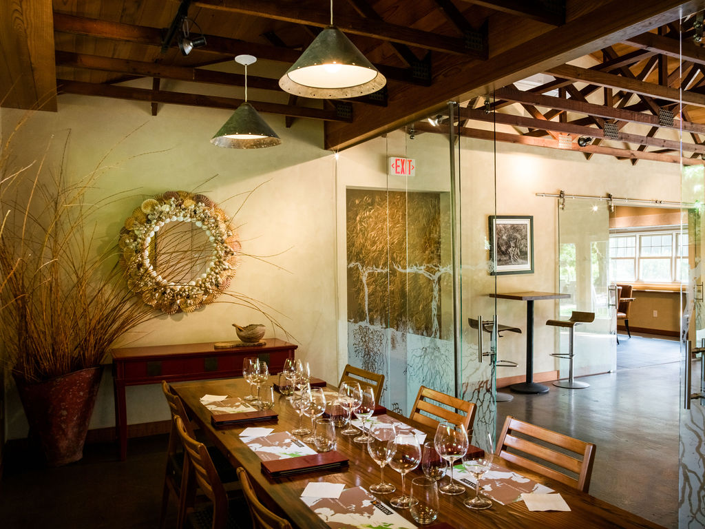 This inviting dining room features a warm, rustic aesthetic with exposed wooden ceiling trusses and a large, rich wood-toned dining table. A decorative seashell-encrusted circular mirror serves as a focal point on the wall, while a glass-walled partition creates an elegant transition to an adjoining lounge area. The space feels sophisticated yet earthy, with thoughtful ambient lighting and a professional, welcoming atmosphere suitable for dining or intimate events.