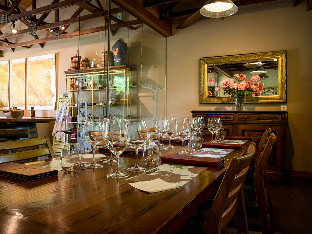 This inviting dining room space is set up for a wine tasting or formal event, featuring a large, polished wood table surrounded by wooden chairs. The room is accented by a decorative mirror reflecting a vase of flowers, a classic dark wood sideboard, and a glass-enclosed display case, creating an elegant and sophisticated atmosphere.
