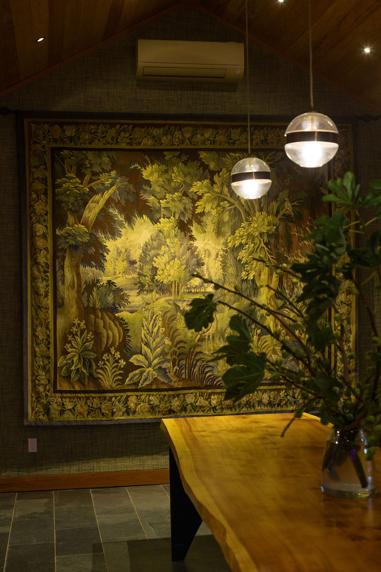 This warm and inviting dining space features a live-edge wooden table in the foreground and a large, intricate landscape tapestry adorning the wall. The room is accented by contemporary spherical pendant lighting, a vaulted wood-paneled ceiling, and dark slate floor tiling. The sophisticated decor creates a rich, textured atmosphere suitable for an upscale dining area.