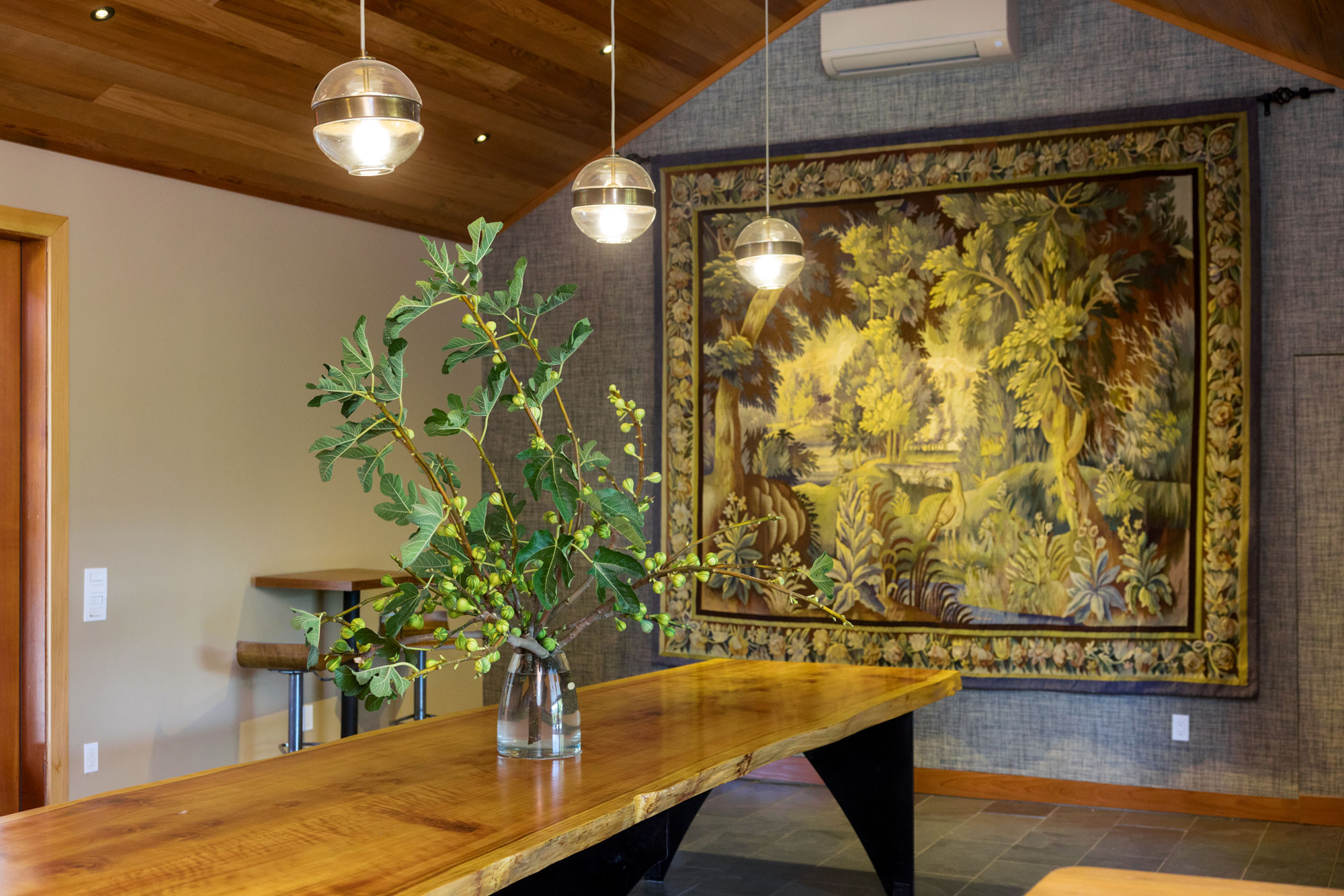 This interior space features a dramatic, live-edge wood table as its centerpiece, topped with a striking fig branch arrangement in a glass vase. A large, ornate landscape tapestry hangs on the textured grey accent wall, complemented by modern, globe-style pendant lights suspended from a warm wood-paneled vaulted ceiling. The space conveys a refined, rustic-meets-contemporary aesthetic suitable for a high-end dining or gathering area.
