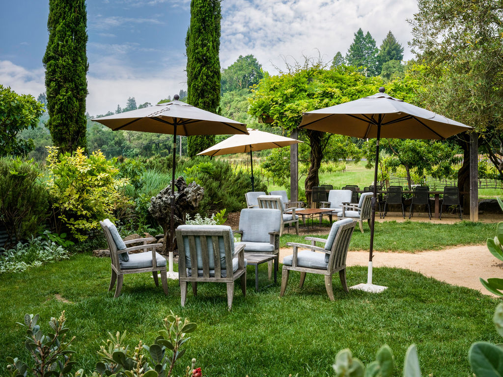 This serene garden setting features clusters of comfortable outdoor seating arranged under large, neutral-toned patio umbrellas on a lush green lawn. The space is complemented by mature trees, including tall cypresses, and offers a peaceful, well-manicured area for relaxation. It provides an inviting atmosphere that suggests a premium outdoor living or event space typical of a high-end estate or vineyard.