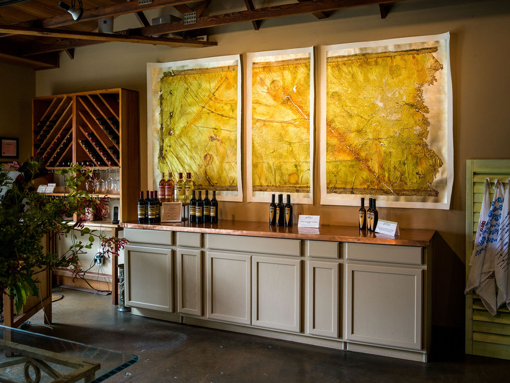This wine tasting room features a copper-topped counter showcasing various bottled wines, set against a wall adorned with three large, textured, golden-hued abstract artworks. To the left, a built-in wooden wine rack provides storage, while the overall aesthetic is warm and inviting with rustic wood ceiling beams. The space creates an upscale, comfortable atmosphere ideal for wine display and appreciation.