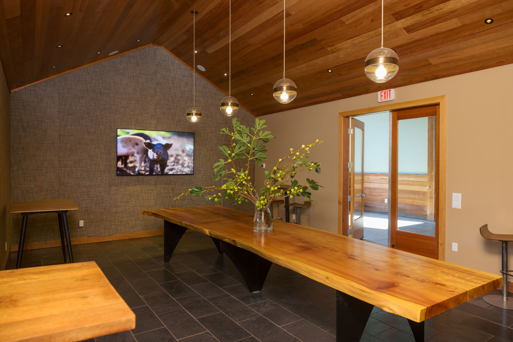 This modern, rustic-inspired interior space features a dramatic vaulted wood-plank ceiling and a large, live-edge wood table that serves as a central focal point. The room includes stylish pendant lighting, textured grey walls, and a large flat-screen display, creating an inviting atmosphere for social gatherings or meetings. A wide doorway leads to an adjacent space, emphasizing an open and airy design suitable for a community building or event area.