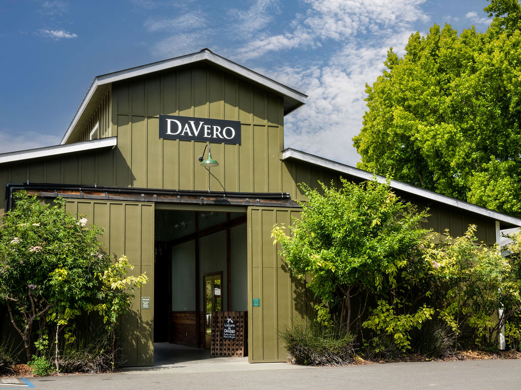 This image showcases the welcoming front entryway of the DaVero winery building, featuring a rustic-modern barn aesthetic with vertical green siding and large sliding barn-style doors. The inviting open doorway reveals a glimpse of the interior, while lush greenery and a charming 'Welcome to DaVero' sign add to the property's appealing and tranquil atmosphere.