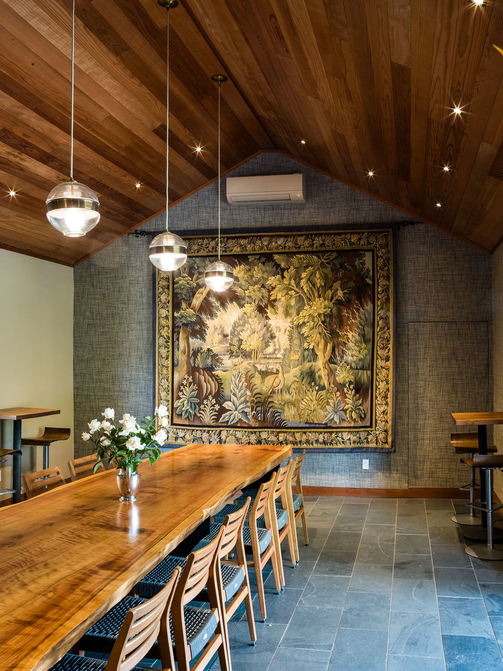 This sophisticated dining area features a stunning live-edge wooden table as its centerpiece, complemented by a rustic tapestry hanging on a textured grey wall. The vaulted wood-plank ceiling, adorned with recessed and globe pendant lighting, adds architectural character and warmth to the space. The stone tile flooring and integrated bar seating areas complete this elegant room, which is perfect for refined entertaining.