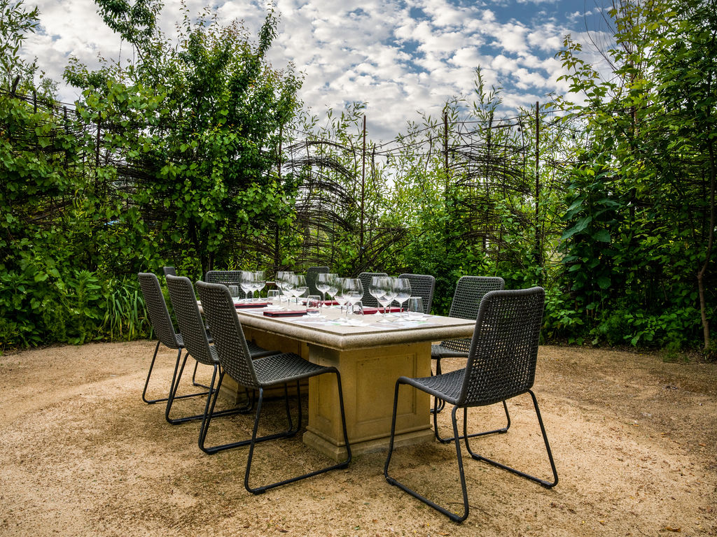 This outdoor dining space features a rectangular stone table set with glassware, surrounded by modern dark woven chairs. It is situated on a light-colored gravel ground, enclosed by a natural woven willow-style privacy screen and lush green foliage. The arrangement creates an inviting and serene al fresco dining atmosphere perfect for entertaining.