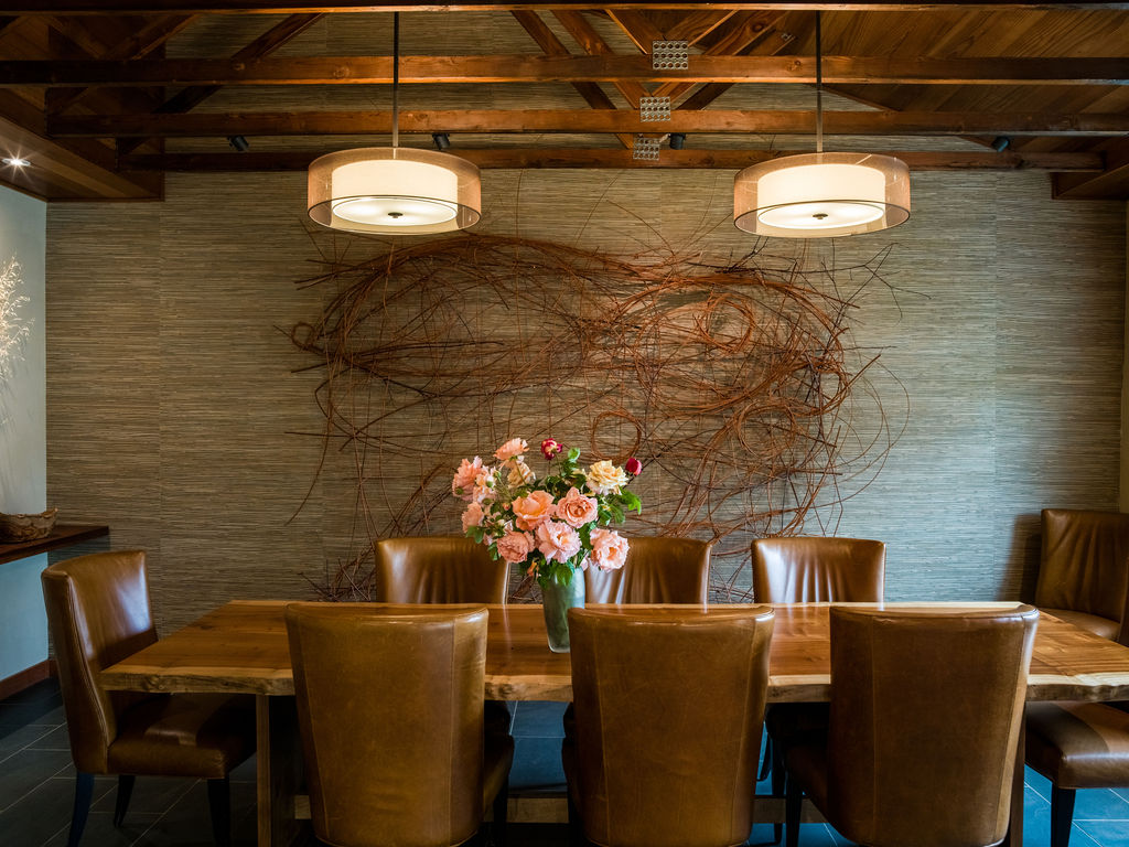 This elegant dining room features a rustic live-edge wood dining table surrounded by high-backed tan leather chairs, set against a textured accent wall adorned with an intricate decorative branch installation. The space is illuminated by two sophisticated dual-tiered drum pendant lights, and the exposed wooden beam ceiling adds a layer of warmth and architectural character. A vibrant bouquet of roses serves as a refined centerpiece, creating an inviting atmosphere for formal or casual dining.