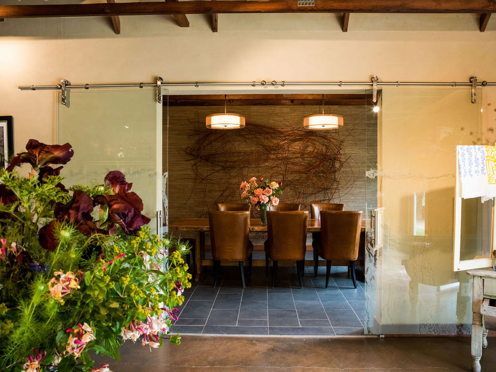 This elegant dining room is framed by large, sliding glass barn-style doors that provide a modern transition from the foreground space. The room features a rustic-chic aesthetic with a live-edge wooden table, leather-upholstered chairs, and unique wall art consisting of intertwined branches against a textured backdrop. Dual pendant lights and slate flooring enhance the sophisticated, warm atmosphere of this well-curated dining area.