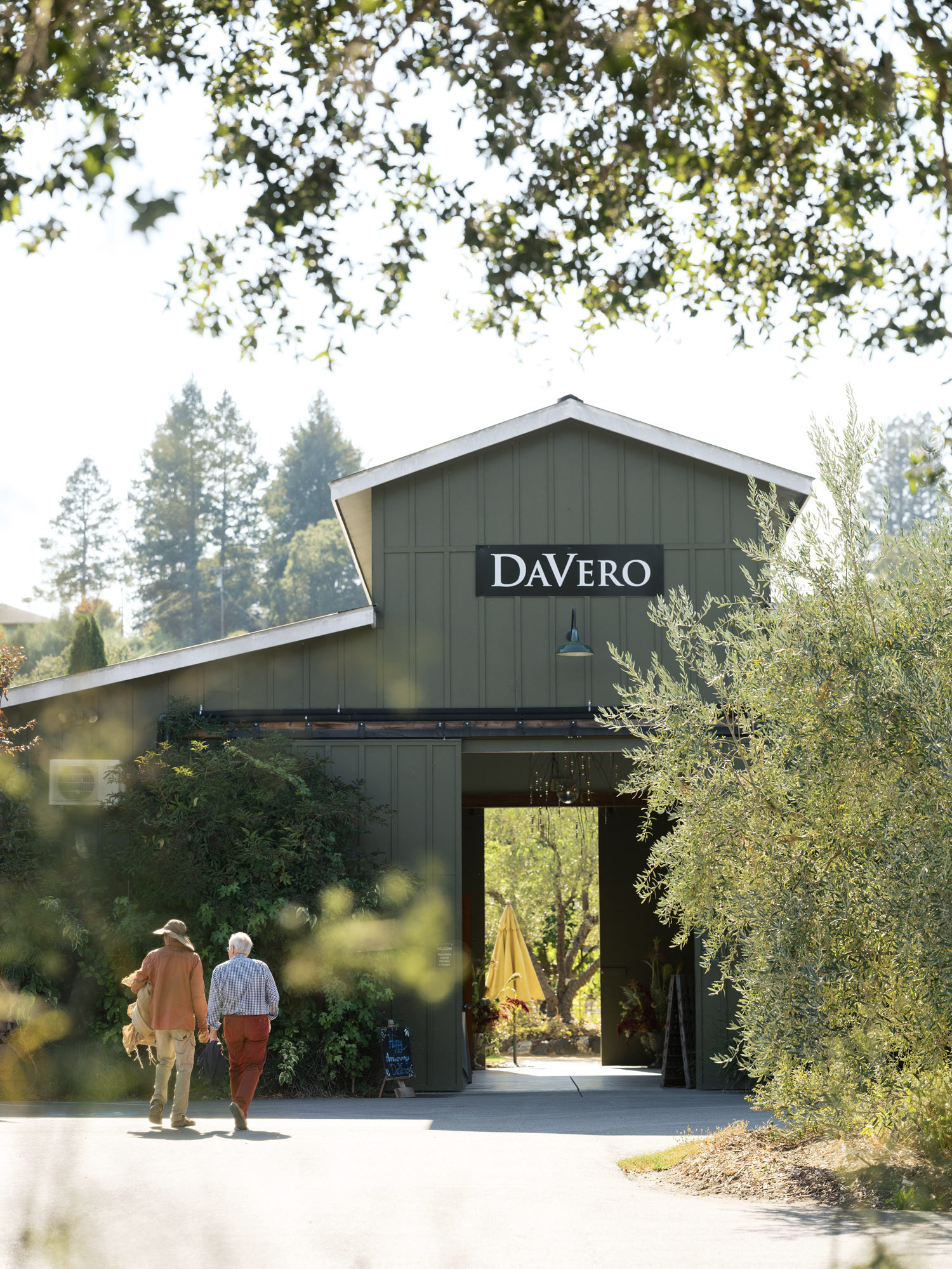This image showcases a rustic, green-sided barn-style structure identified as DaVero, serving as a charming entryway to a vineyard or estate. The building features a large open doorway, a clear sign with the name, and is framed by mature trees and greenery, creating a welcoming and picturesque first impression. Two individuals are walking toward the entrance, highlighting the scale and inviting nature of this pastoral property.