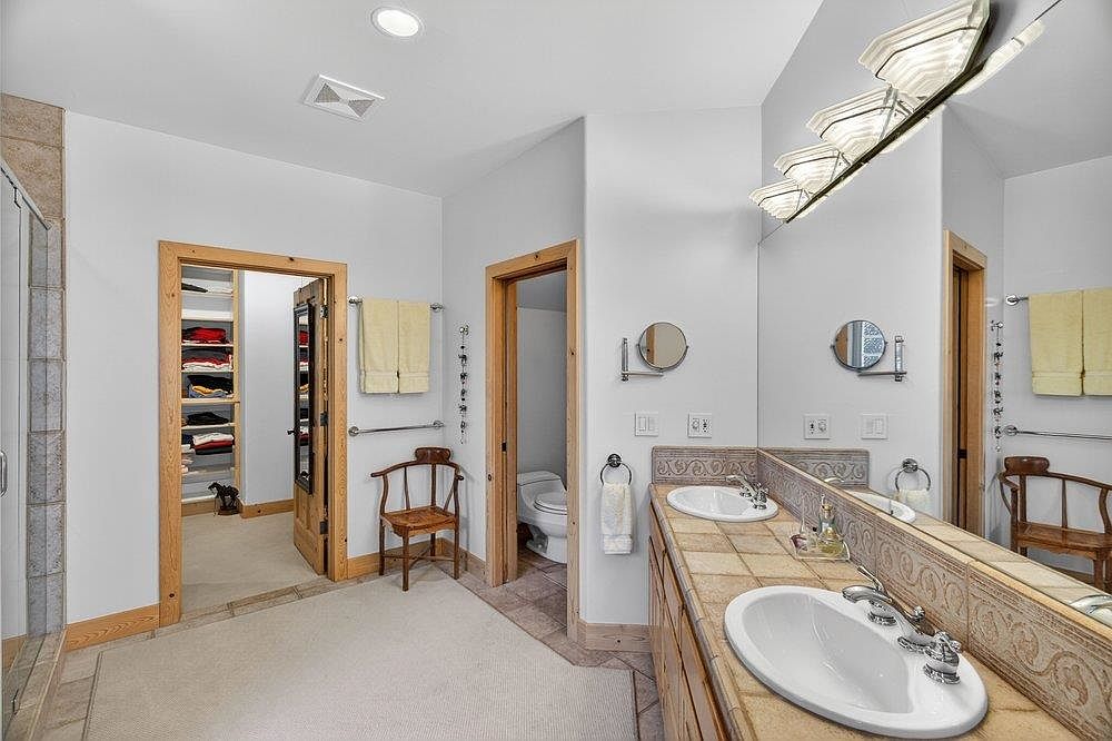 This primary bathroom features a dual-sink vanity with a tiled countertop and a large wall-mounted mirror. The space includes a separate water closet, a walk-in closet with built-in shelving, and a wooden chair, all tied together by neutral wall tones and light wood trim. The perspective captures the functional layout of the room, highlighting the transition between the vanity area, the closet, and the private toilet room.