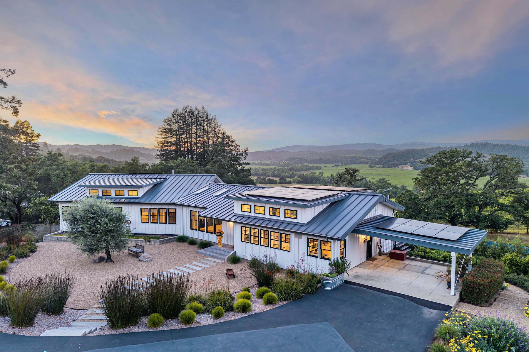 This elevated aerial perspective captures a modern farmhouse-style residence set against a sprawling rural landscape at sunset. The home features a dark metal roof with integrated solar panels, white siding, and numerous windows that reflect the warm evening light. A gravel courtyard with stone pavers leads to the entrance, while a paved driveway extends under a covered carport, all surrounded by lush greenery and rolling hills in the distance.
