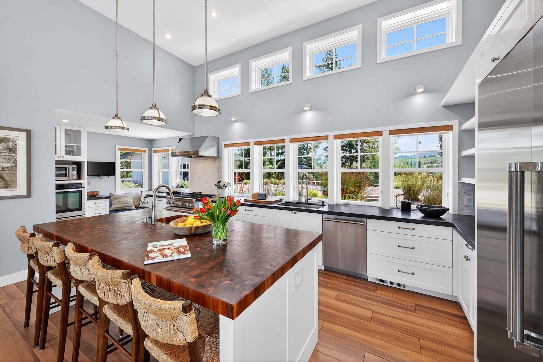 This bright and airy kitchen features a large central island with a dark wood butcher-block countertop and seating for four. The space is defined by white cabinetry, dark countertops, and a series of large windows that provide abundant natural light and views of the surrounding landscape. High ceilings with pendant lighting and a neutral color palette create an inviting, open-concept atmosphere.