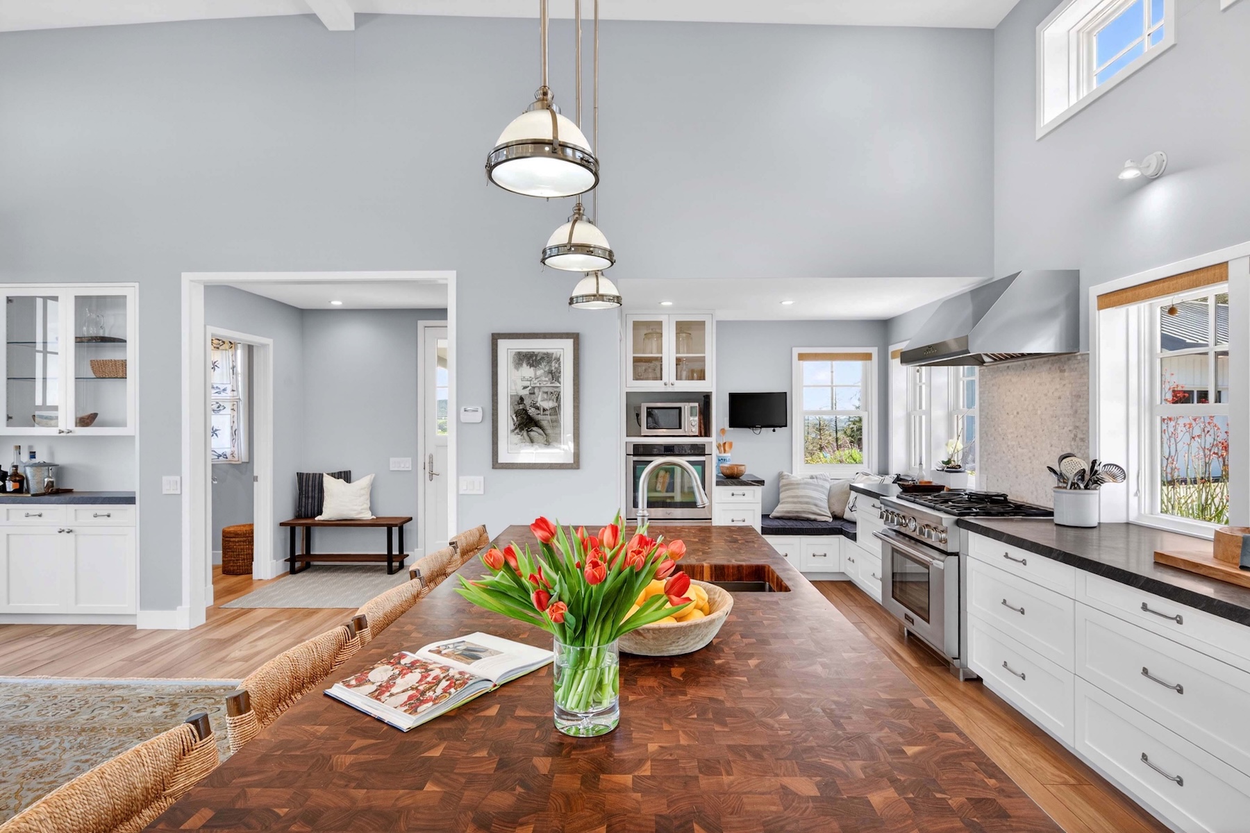 This bright and airy kitchen features a large central island with a dark wood butcher-block countertop, serving as the focal point of the room. White cabinetry, stainless steel appliances, and a professional-style range hood create a clean, modern aesthetic, while a cozy window seat nook adds a touch of charm. The space is illuminated by three pendant lights hanging above the island, and the open-concept layout flows seamlessly into an adjacent hallway with a built-in bench.