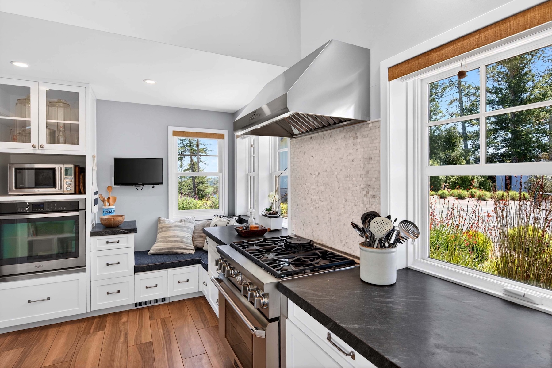 This bright and functional kitchen features white cabinetry, dark stone countertops, and high-end stainless steel appliances, including a professional-style gas range with a matching vent hood. A cozy built-in window seat with patterned cushions provides a charming nook, while large windows allow for ample natural light and views of the surrounding greenery. The space is finished with light-toned wood flooring and a textured tile backsplash, creating a warm and inviting atmosphere.