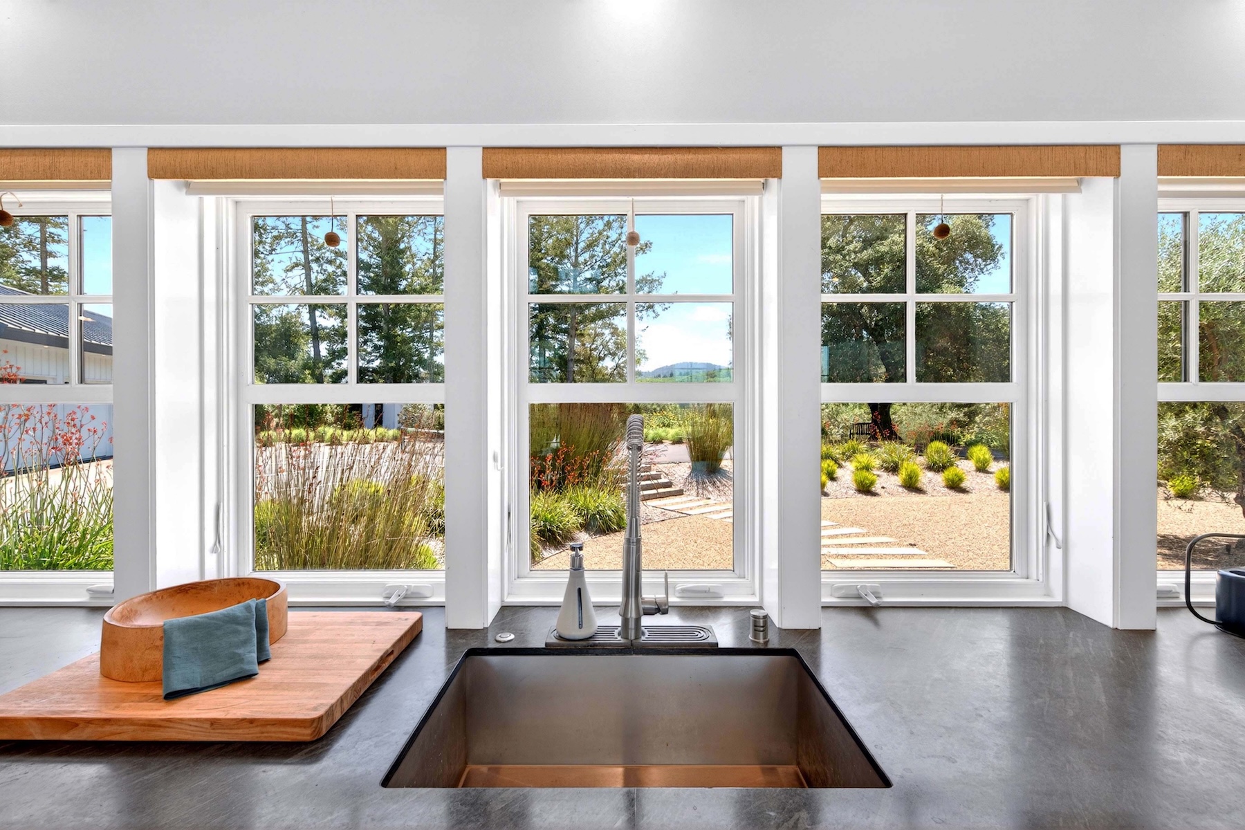 This kitchen features a large, deep stainless steel sink set into a dark, matte-finish countertop. Three large windows provide a bright, symmetrical view of the landscaped garden outside, framed by simple woven shades. A wooden bowl and cutting board sit to the left, creating a warm, functional aesthetic that emphasizes the connection between the indoor workspace and the natural surroundings.