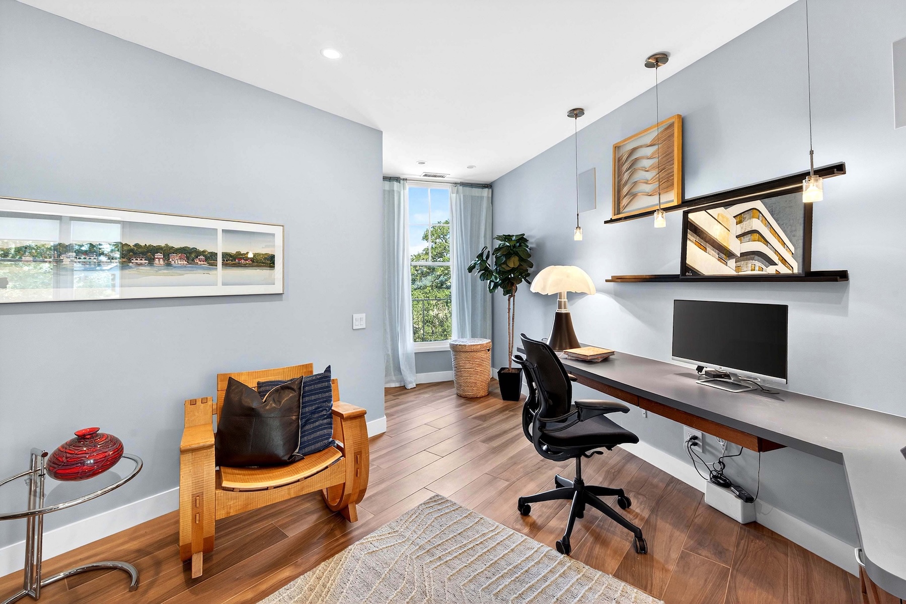 This modern home office features a sleek, wall-mounted desk with a computer monitor and a comfortable ergonomic chair. The room is accented by a unique wooden rocking chair, a large potted plant, and tasteful wall art, all set against light blue walls and warm-toned wood flooring. A large window at the end of the room provides natural light and a view of the outdoors, creating a bright and productive workspace.