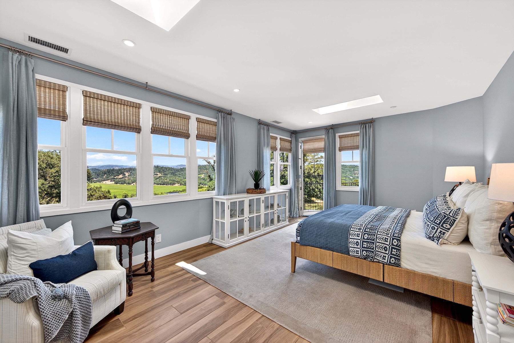 This bright and airy primary bedroom features light blue walls, wood-look flooring, and a large area rug that anchors the space. A bed with a patterned blue and white quilt sits beside a white cabinet, while a cozy armchair and side table are positioned near a wall of windows offering expansive views of the surrounding landscape. The room is illuminated by natural light from the windows and a skylight, creating a serene and inviting atmosphere.