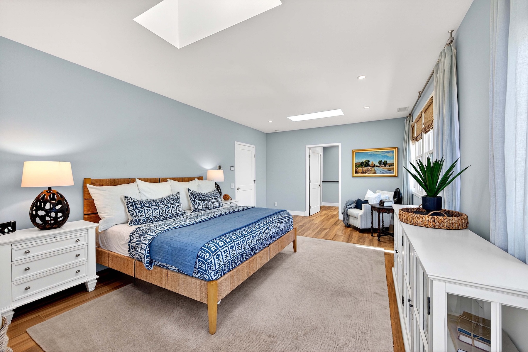 This spacious primary bedroom features light blue walls, warm wood flooring, and a large area rug centered under a king-sized bed with a woven headboard. The room is brightened by two skylights and large windows draped in soft curtains, while a white dresser and a cozy seating area with an armchair add functional elegance. The perspective is a wide-angle shot from the foot of the bed, capturing the airy, tranquil atmosphere of the suite.