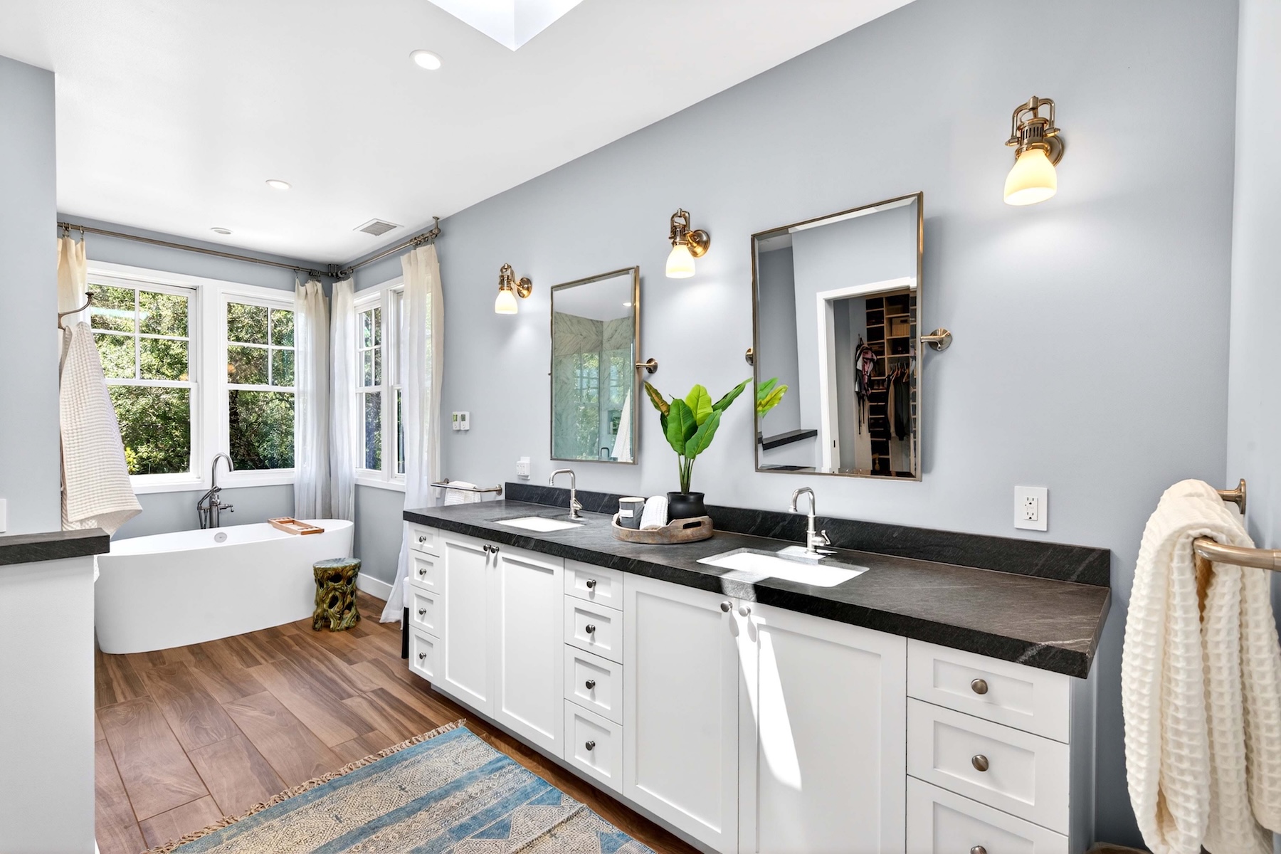 This bright and airy primary bathroom features a double vanity with white cabinetry and a dark stone countertop, complemented by two rectangular mirrors and wall-mounted light fixtures. To the left, a freestanding soaking tub is positioned near large windows that allow for ample natural light, while a patterned area rug adds texture to the wood-look flooring. The space is finished with light blue walls and a clean, modern aesthetic that creates a serene and spa-like atmosphere.