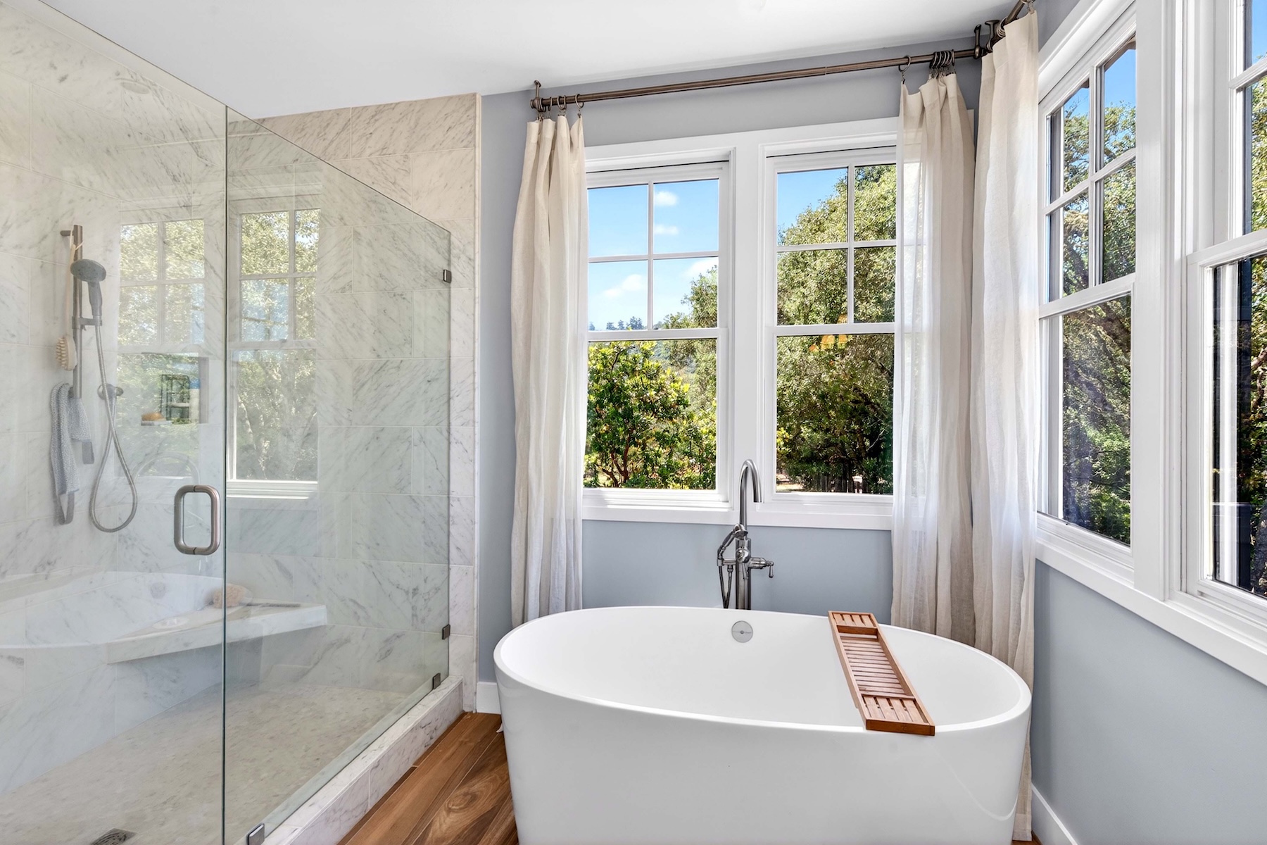 This bright primary bathroom features a freestanding soaking tub positioned beneath large windows that offer a view of the surrounding trees. To the left, a glass-enclosed walk-in shower is finished with light-toned stone tile and includes a handheld showerhead. The space is accented by light wood flooring and soft blue wall paint, creating a serene and spa-like atmosphere.