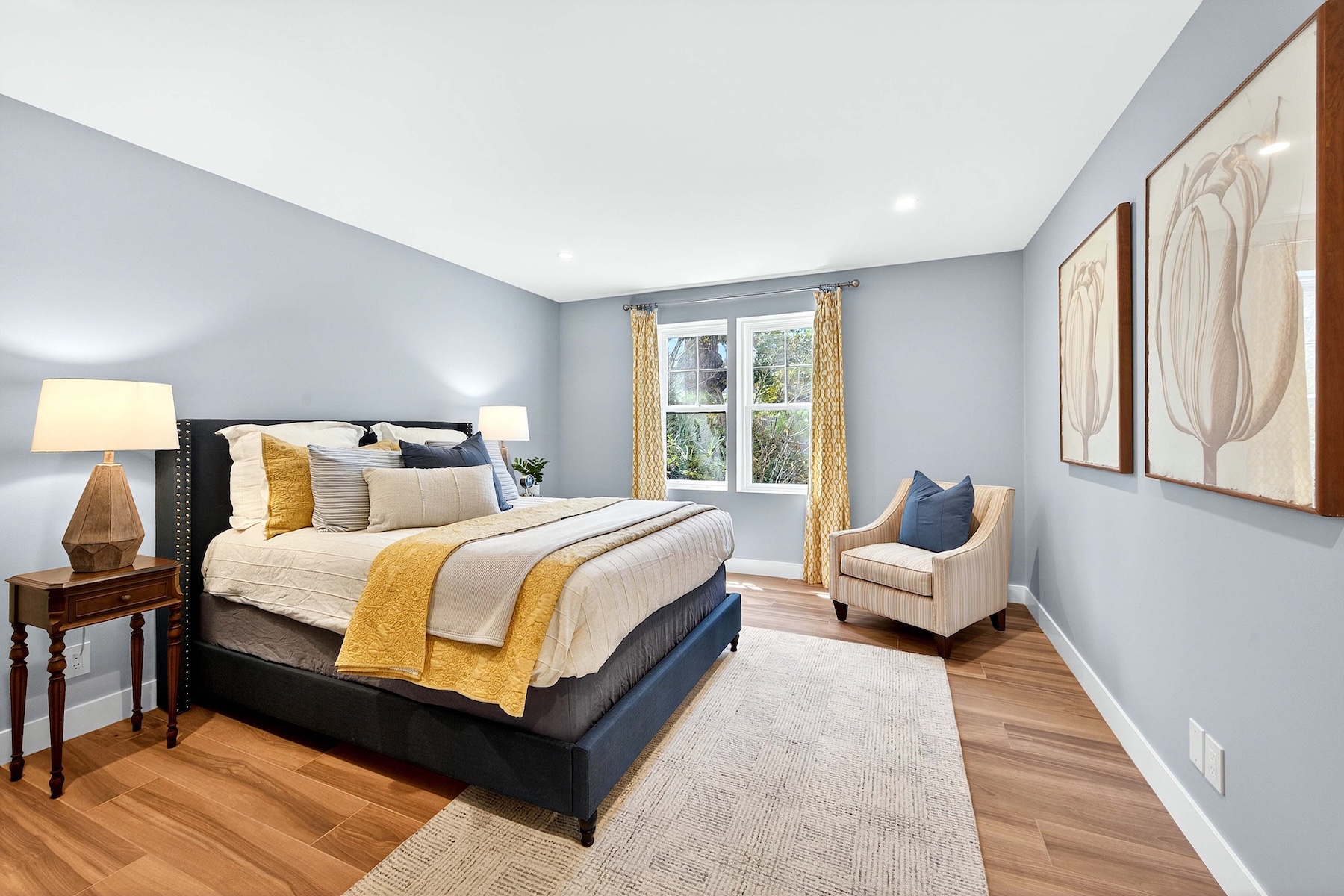 This primary bedroom features a neutral color palette with soft blue walls and warm wood-look flooring. A dark upholstered bed serves as the focal point, accented by yellow and blue throw pillows, while a striped armchair sits in the corner near large windows dressed in patterned yellow curtains. Two framed botanical art pieces hang on the right wall, creating a serene and well-appointed atmosphere.