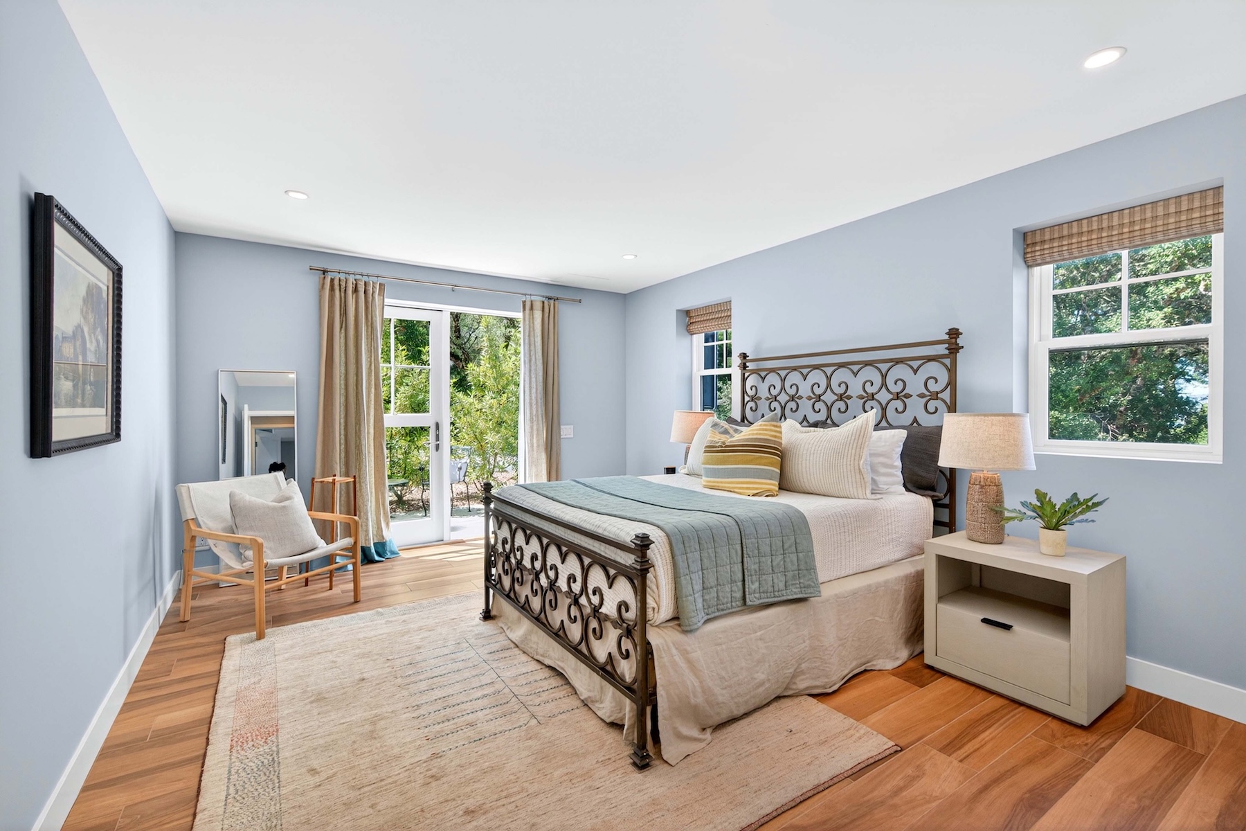This bright and airy primary bedroom features light blue walls, warm wood-toned flooring, and a large area rug that anchors the space. A metal-framed bed serves as the focal point, complemented by a bedside table with a textured lamp and a cozy seating area near the glass door that leads to the outdoors. The room is filled with natural light, creating a serene and inviting atmosphere.