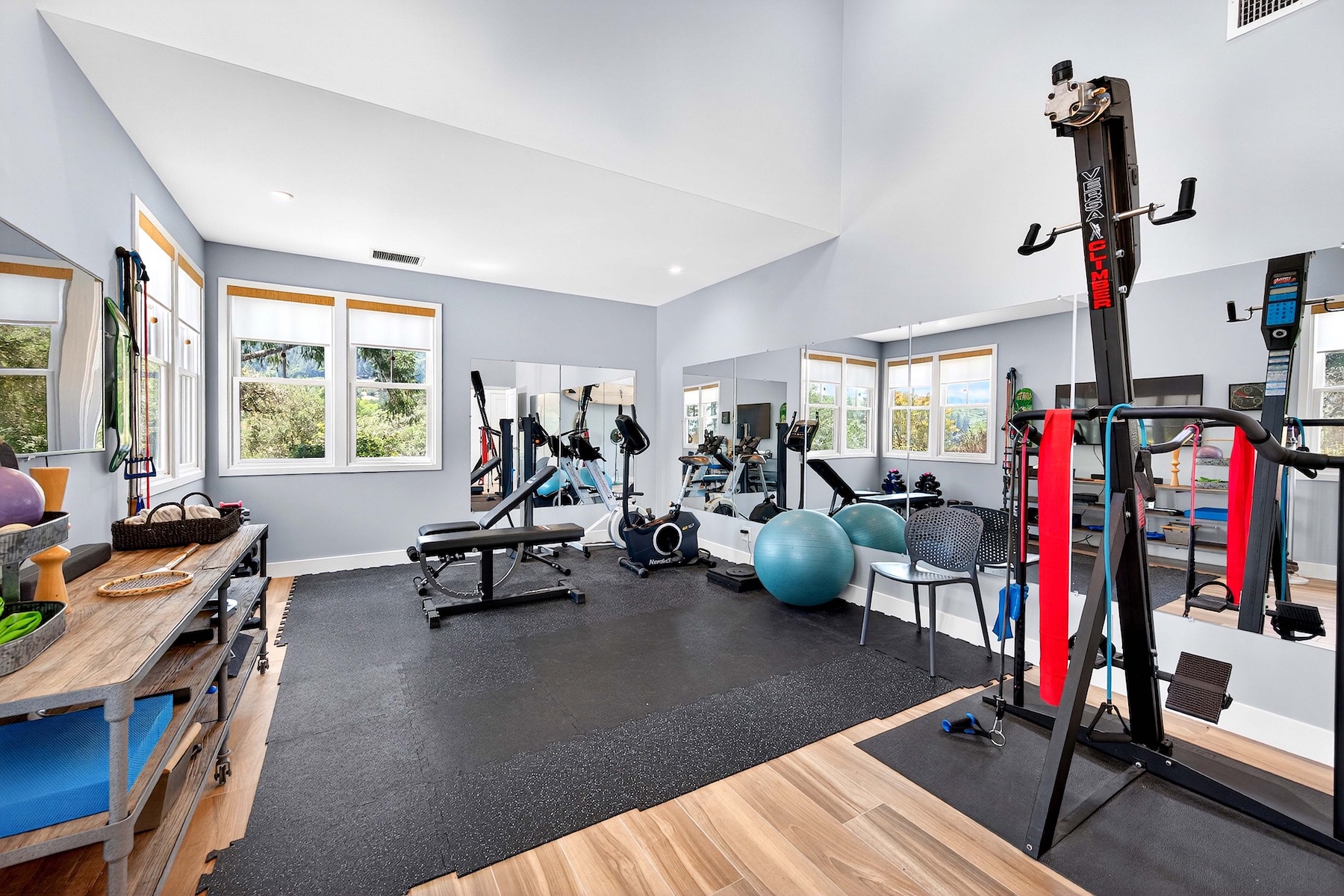 This home gym features a spacious layout with light gray walls, high ceilings, and large windows that provide ample natural light. The room is equipped with a variety of fitness equipment, including a VersaClimber, a weight bench, a stationary bike, and exercise balls, all set upon a durable black rubber mat. Large wall-to-wall mirrors enhance the sense of space, creating a bright and functional workout environment.