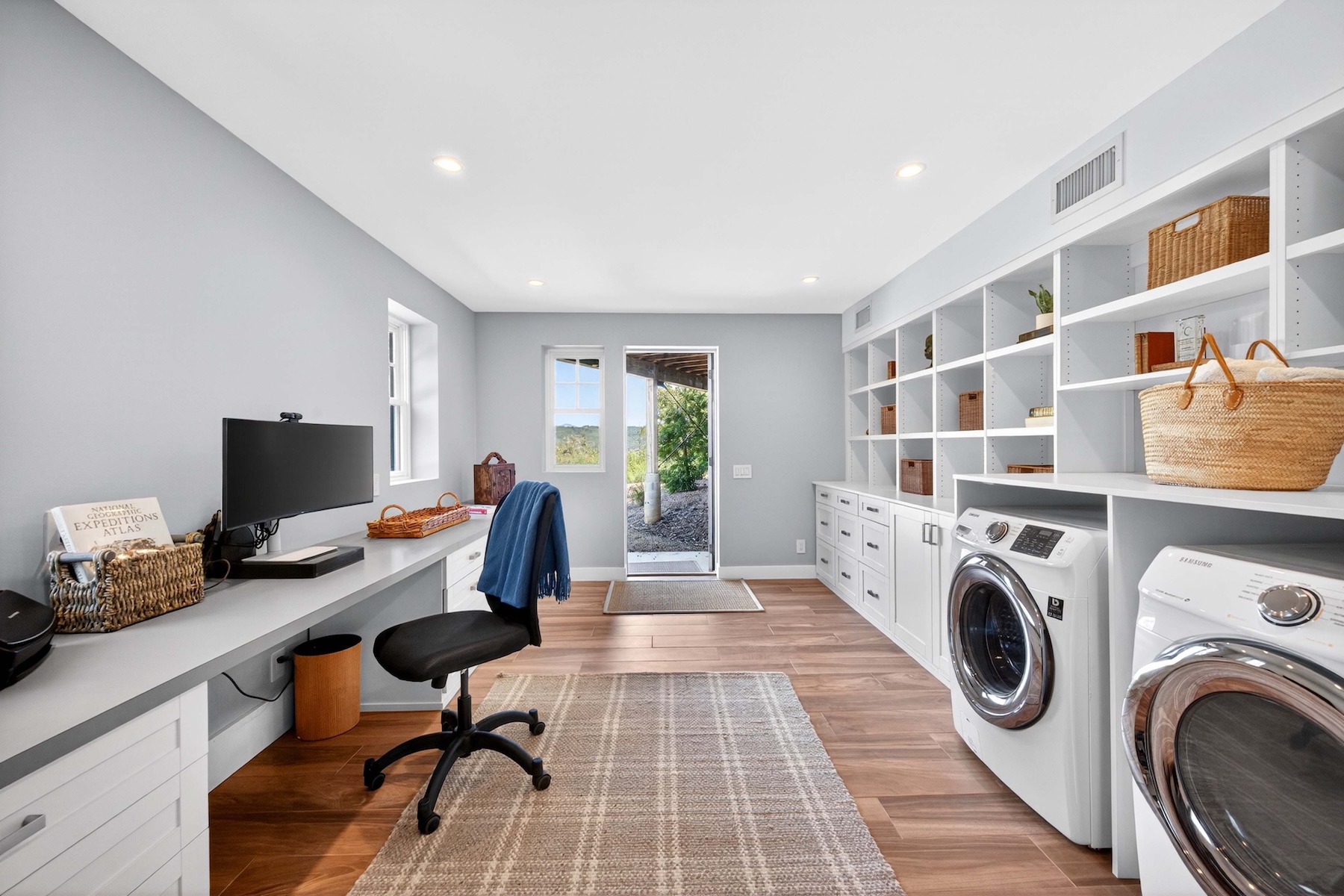 This versatile space functions as both a laundry room and a home office, featuring a long desk area with a computer monitor and a built-in shelving unit. The room is finished with light-toned walls, wood-look flooring, and white cabinetry that houses a side-by-side washer and dryer set. A door at the far end provides direct access to the outdoors, creating a bright and functional multi-purpose environment.