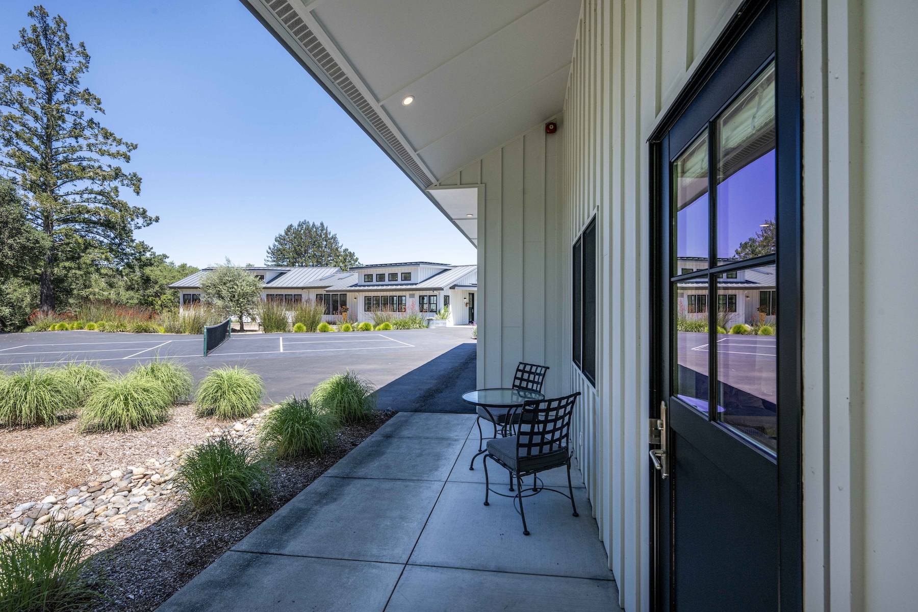 This image captures a covered concrete patio area featuring a small bistro table and two metal chairs, positioned next to a dark-framed glass door. The perspective looks out from the porch toward a paved area that serves as a tennis court, set against a backdrop of lush green trees and a clear blue sky. The architecture features light-colored vertical siding, creating a clean, modern aesthetic that blends seamlessly with the surrounding outdoor space.