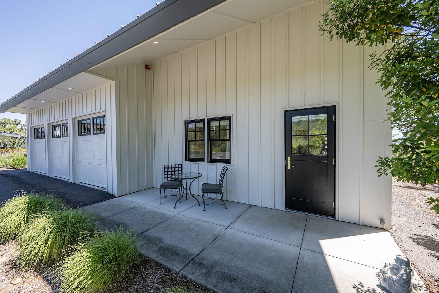 This exterior view captures a modern, farmhouse-style building featuring white vertical siding and a sleek, dark-trimmed garage section. A concrete patio area in the foreground is furnished with a small bistro set, creating a cozy outdoor seating space adjacent to a black-framed glass door. The scene is bathed in bright, natural sunlight, highlighting the clean lines of the architecture and the well-maintained landscaping.