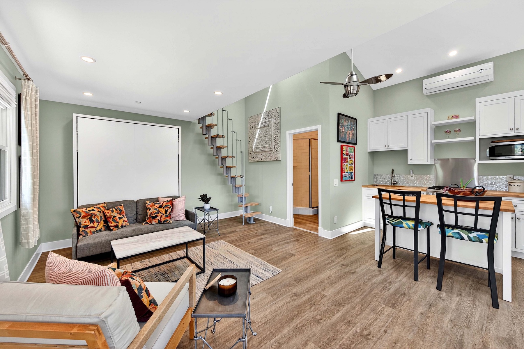 This open-concept living space features a light green color palette, wood-look flooring, and a vaulted ceiling that creates an airy, spacious feel. The room includes a cozy seating area with a grey sofa and patterned accent pillows, a modern kitchen with white cabinetry and a breakfast bar, and a unique floating staircase leading to an upper level. A large white wall-mounted unit serves as a focal point, while the overall design blends contemporary functionality with a bright, inviting aesthetic.