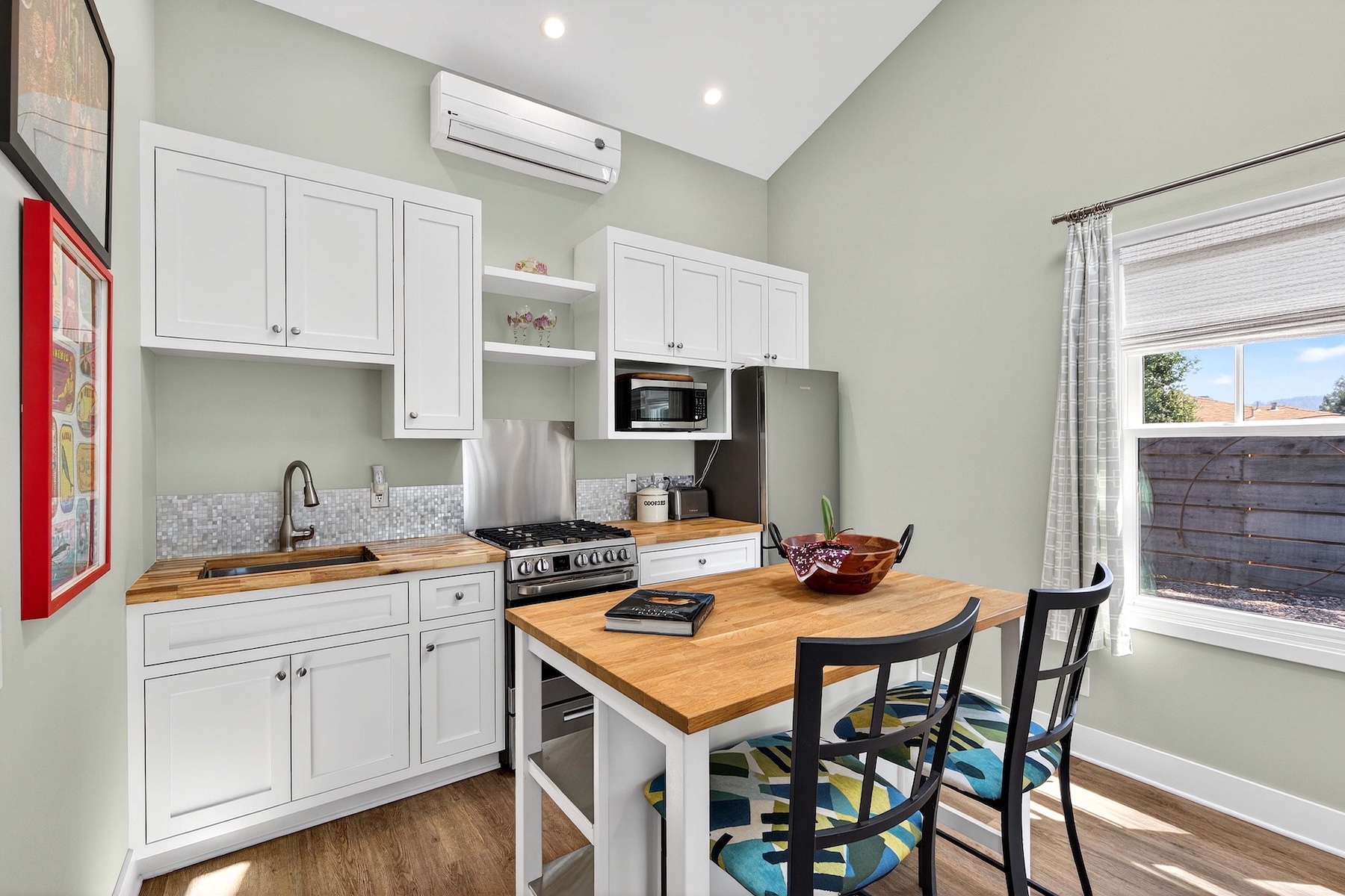 This compact, efficient kitchen features white cabinetry paired with light wood countertops and a mosaic tile backsplash. A central island with matching wood top provides additional workspace and seating with two patterned chairs, while a stainless steel range and refrigerator complete the appliance suite. The space is brightened by recessed lighting and a window with light curtains, creating a clean and functional atmosphere.