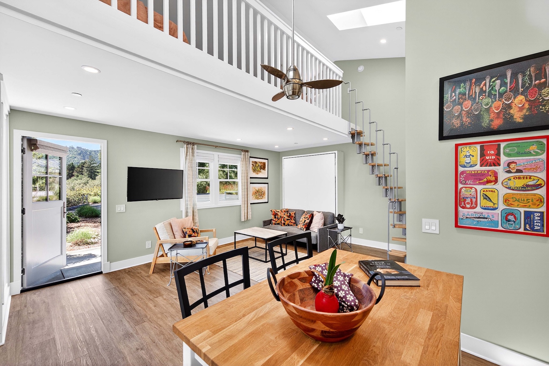 This bright and airy living space features a lofted ceiling with a ceiling fan and a staircase leading to an upper level. The room includes a comfortable seating area with a sofa and armchairs, a wall-mounted television, and a dining area in the foreground with a wooden table. Large windows and an open door provide natural light and a view of the surrounding landscape, creating an inviting and open-concept atmosphere.