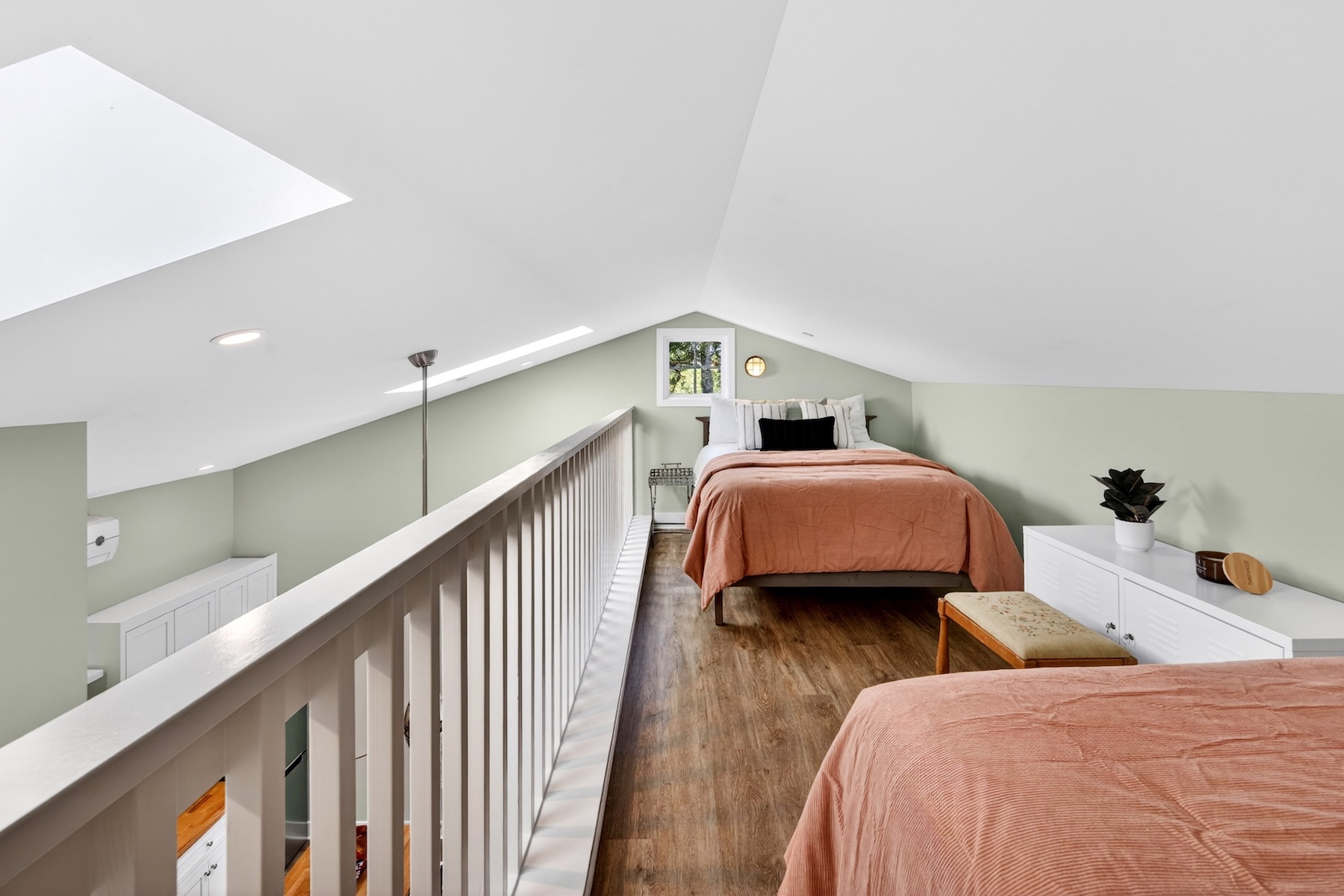 This attic bedroom features a vaulted ceiling with a skylight, creating an airy and open atmosphere. The space is furnished with two beds dressed in warm-toned bedding, set against soft green walls and wood-look flooring. A white railing overlooks the lower level, emphasizing the loft-style layout of this cozy, multi-purpose living area.