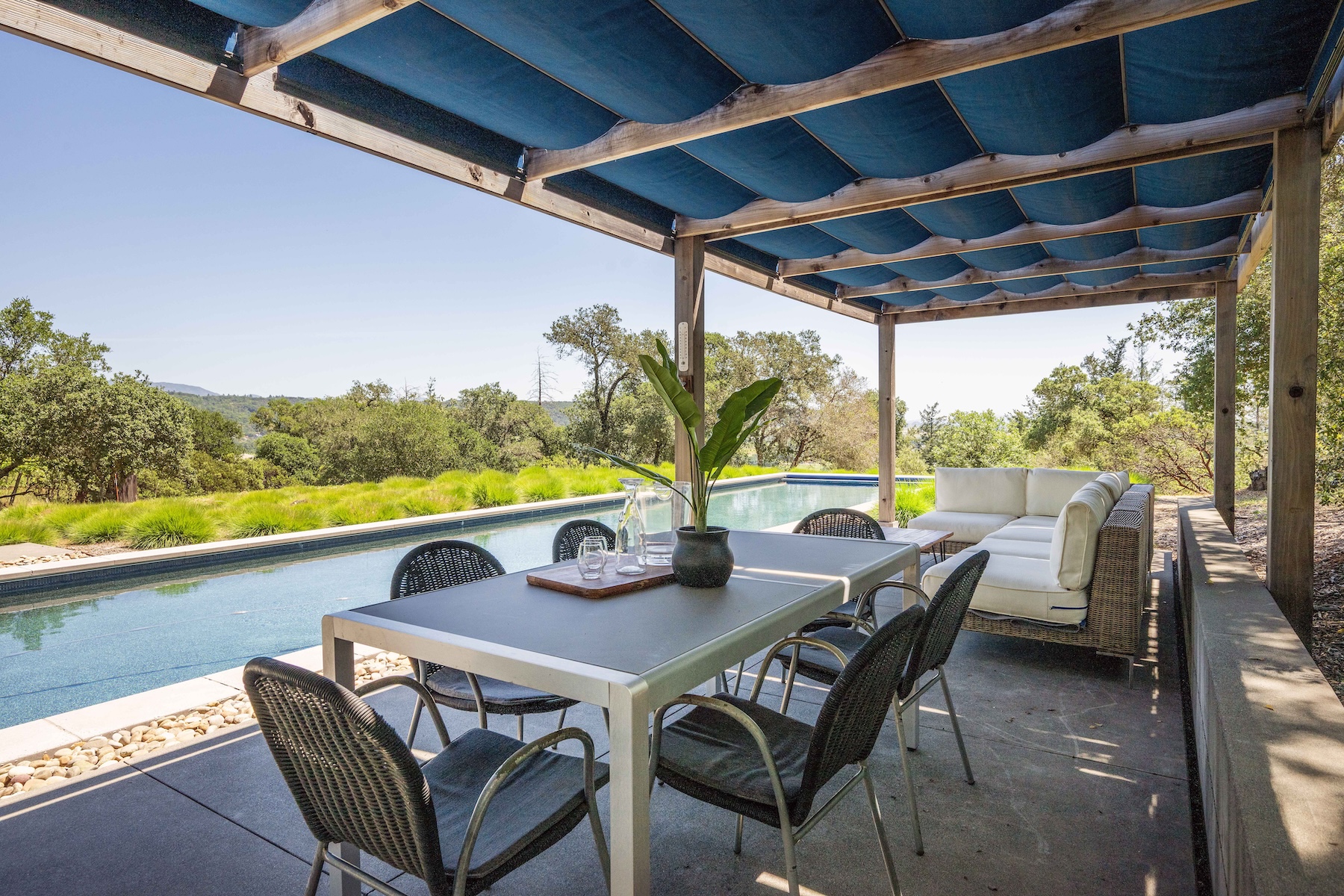 This inviting outdoor living space features a covered patio area with a dining set and a comfortable sectional sofa, perfect for al fresco entertaining. The space is shaded by a wooden pergola with a blue fabric canopy and overlooks a long, narrow lap pool set against a backdrop of lush green trees and rolling hills. The clean, modern design emphasizes a seamless transition between the shaded lounge area and the sunny pool deck.