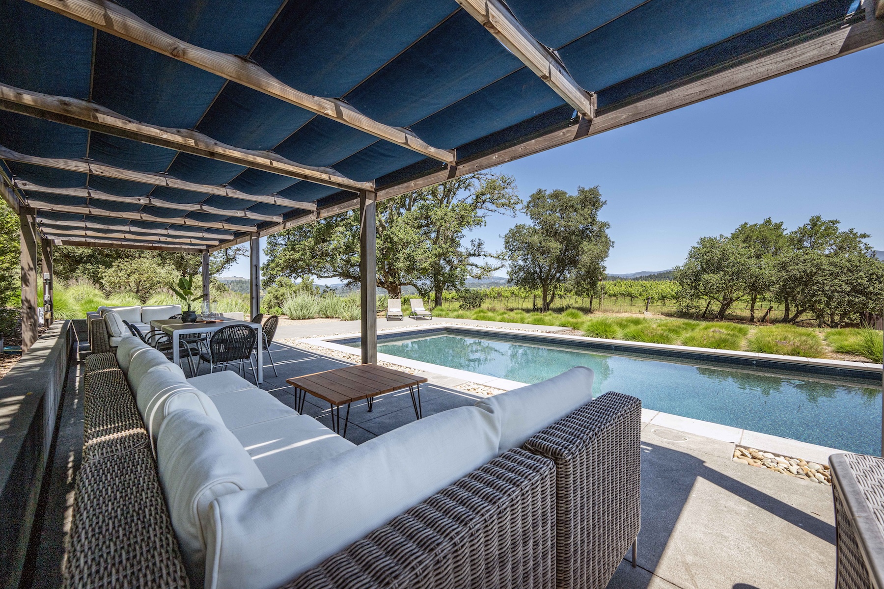 This inviting outdoor living space features a covered patio with a large, L-shaped wicker sectional sofa and a dining area, all shaded by a blue fabric canopy. The patio transitions seamlessly to a rectangular swimming pool, which is set against a backdrop of lush green trees and a vineyard. The scene captures a serene, resort-like atmosphere perfect for relaxation and entertaining.